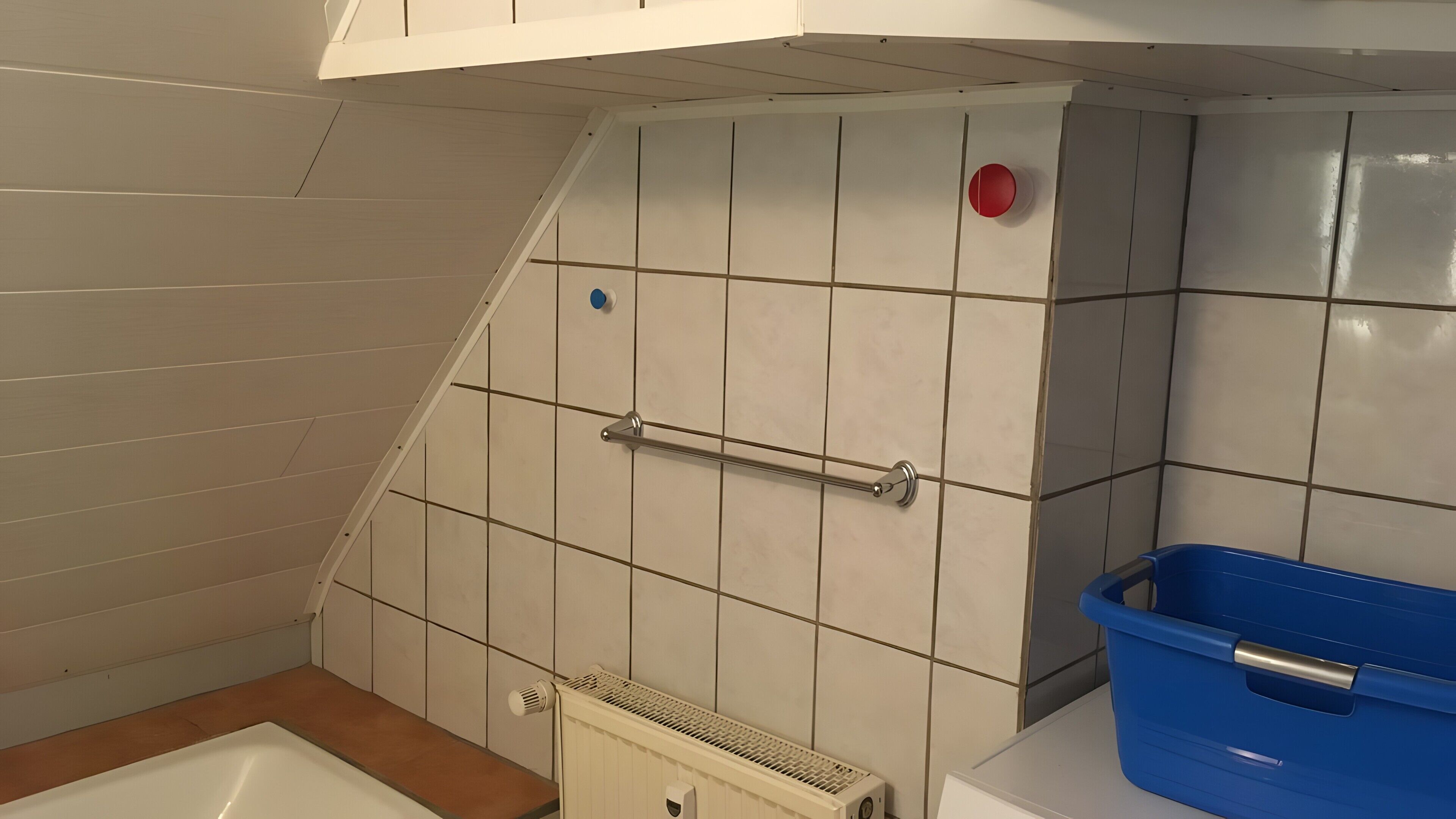 A bathroom with white tiled walls, a white bathtub, a blue trash bin, and a red emergency light.