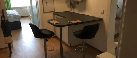 A compact kitchen with a small dining table and two bar stools, a refrigerator, and a window with curtains.