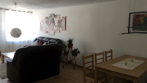 A living room with a sofa, a dining table with chairs, a potted plant, and abstract wall art.