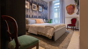 Ett sovrum med en säng, en stol, ett stort fönster och en vägg med inramade tavlor.