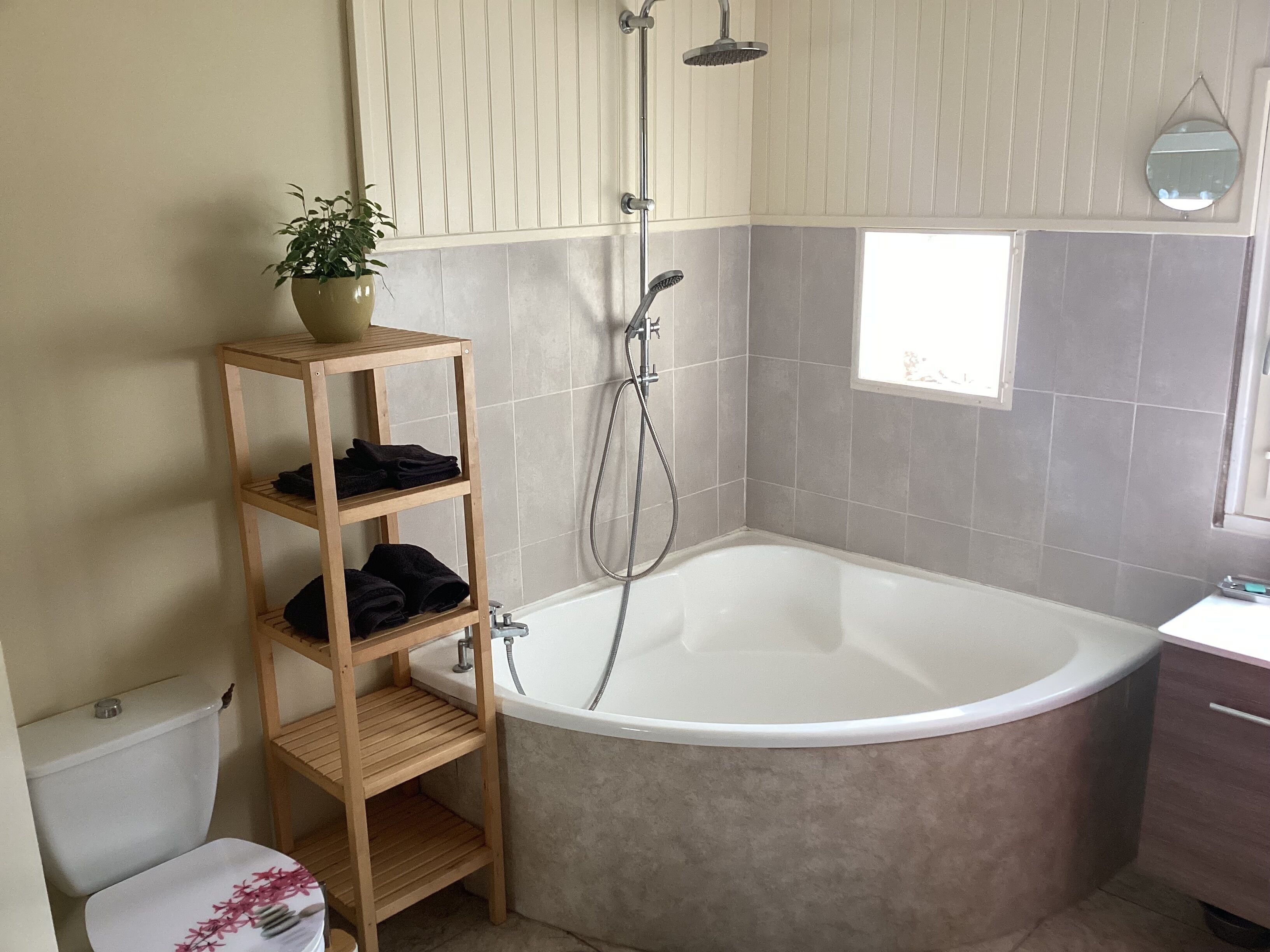 Un baño con bañera, un estante de madera con toallas, una planta en maceta, ducha y ventana.
