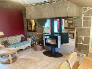 A cozy living room with a stone fireplace, a sofa, a wicker chair, and a small table. - Charming vacation home near Mont-Saint-Michel in the countryside, centrally located (Brécey)