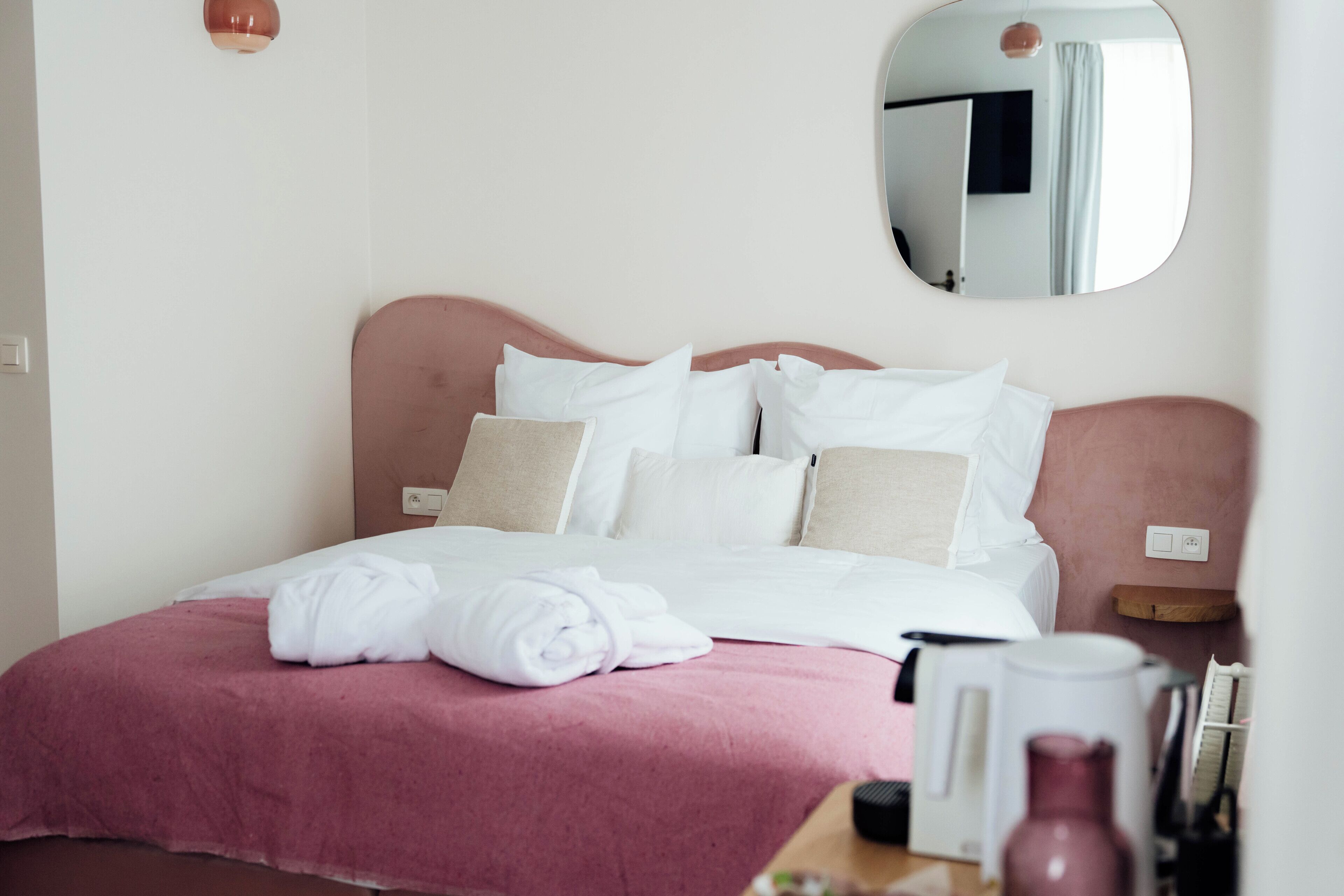 A neatly made bed with white and pink bedding, a bedside table with a lamp, and a mirror on the wall.