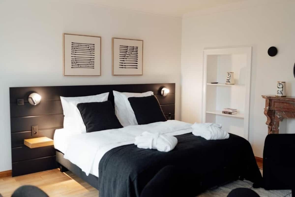 A modern hotel room with a black headboard, white bedding, a bedside table with a lamp, and two framed artworks on the wall.