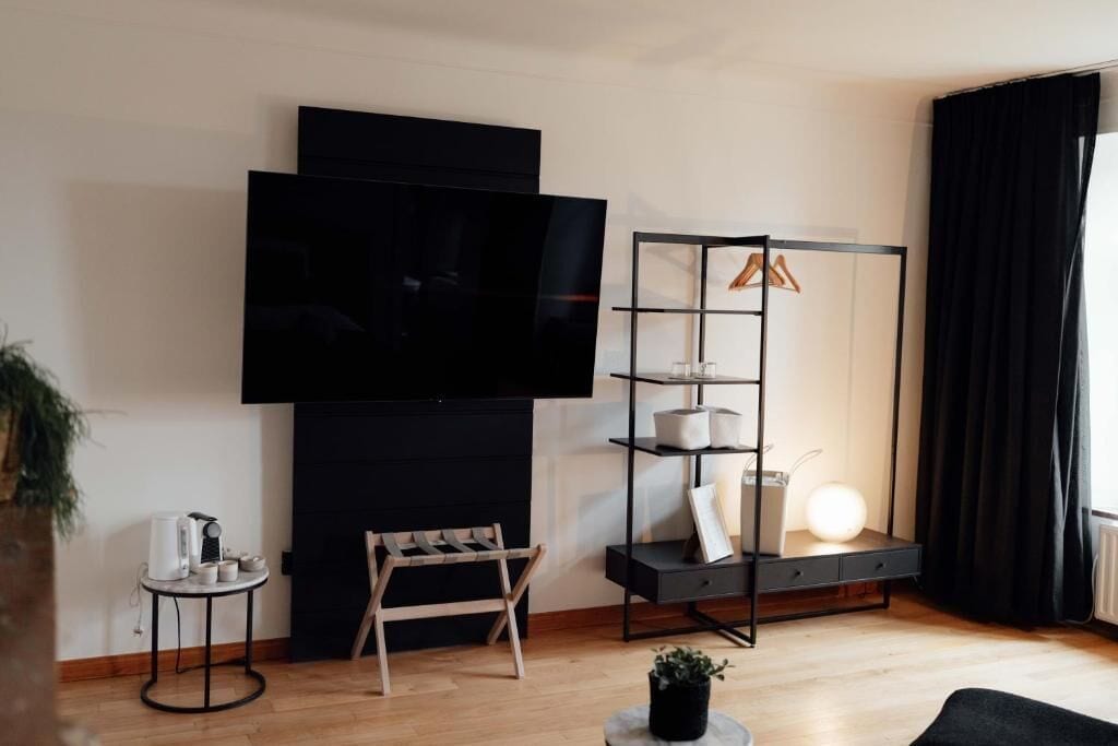 Un salon moderne avec un téléviseur à écran plat, une étagère noire, une petite table avec une bouilloire et un fauteuil.