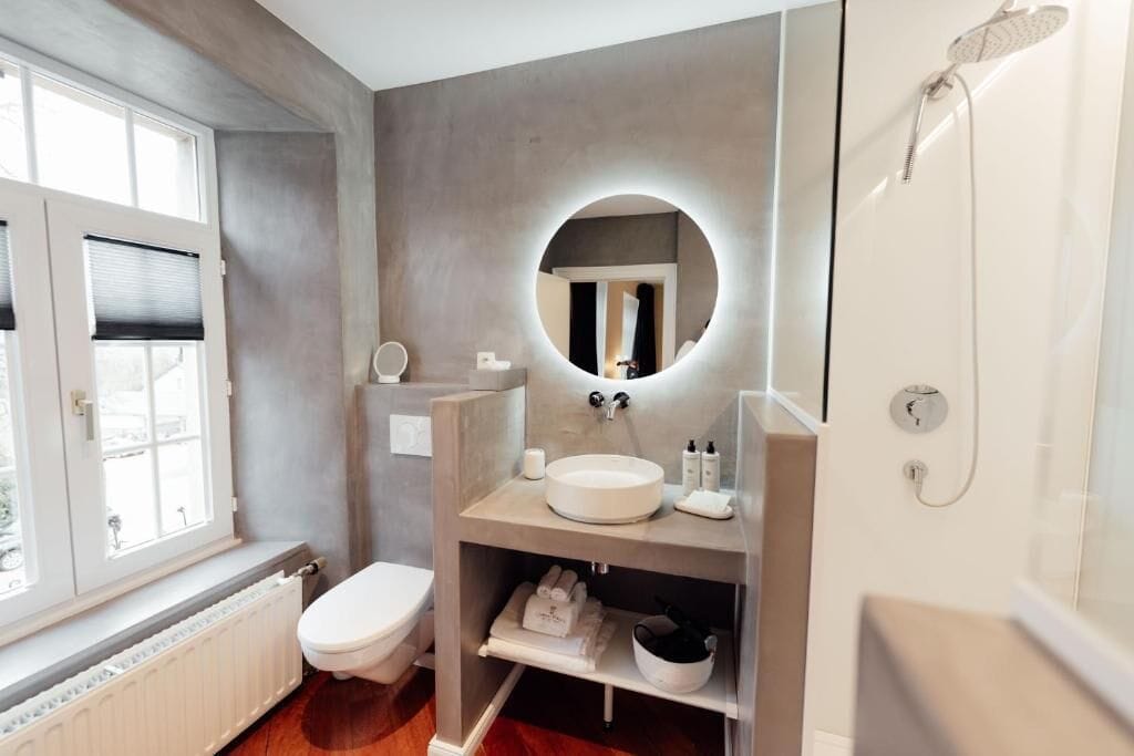 Une salle de bain moderne avec un grand miroir rond, des toilettes, un lavabo et une douche.