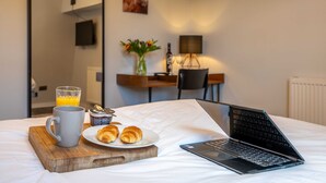 Habitación de hotel con cama, laptop, plato de croissants, taza y vaso de jugo de naranja.