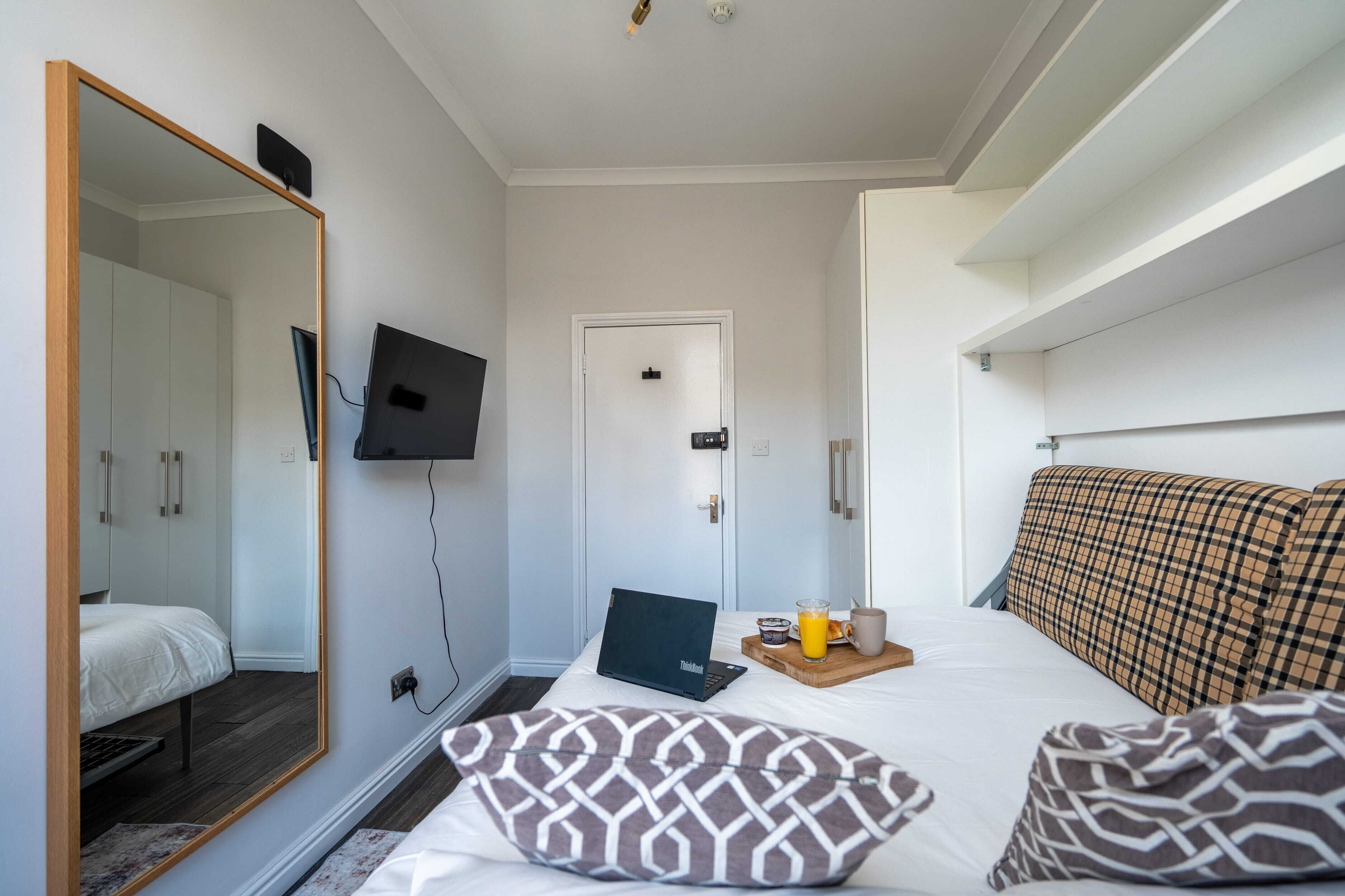 A bedroom with a bed, a laptop, a mirror, a TV, and a door.