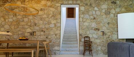 A stone wall, a wooden bench, a dining table with a fruit bowl, a staircase, and a grey sofa.