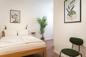 A neatly made bed with white linens, a green potted plant, and two framed artworks on the wall.
