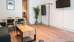 En moderne stue med et træbord, grønne stole, en potteplante, et fladskærms-tv monteret på væggen og en trappe til et andet rum.