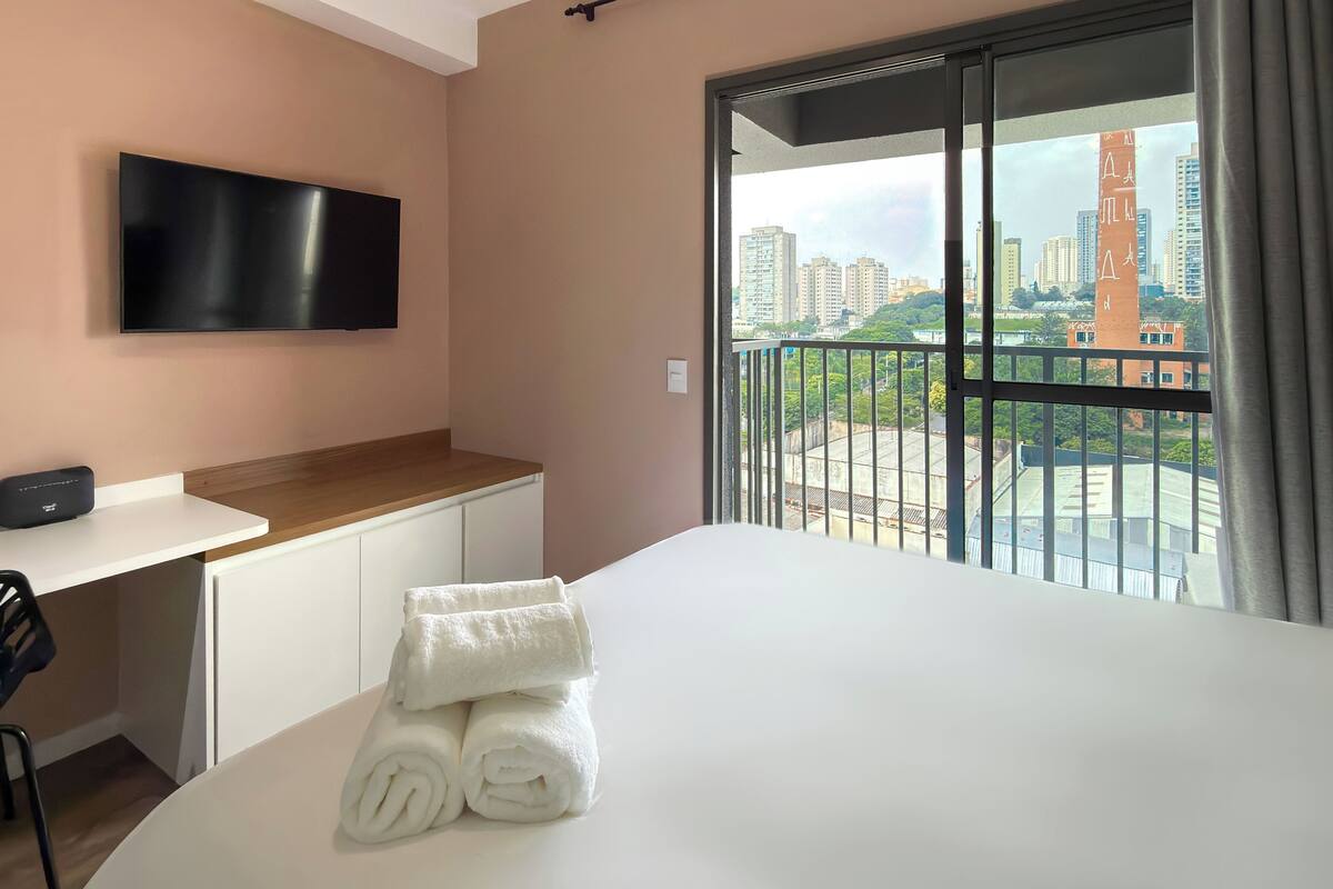 A hotel room with a bed, a TV mounted on the wall, a desk with a chair, and a balcony with a city view.