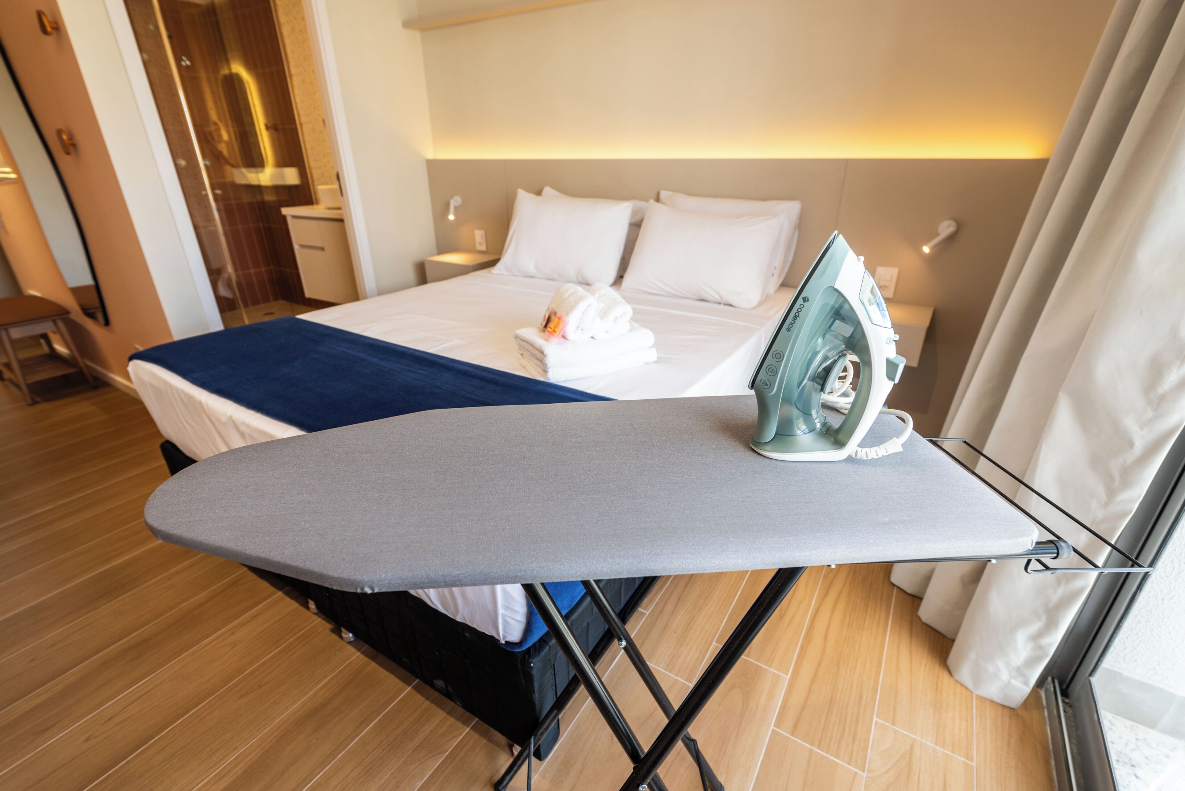 Una cama bien hecha con una manta azul, una tabla de planchar con un planchador y un piso de madera.