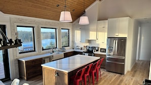 Une cuisine moderne dotée d’un grand îlot central, de meubles en bois, d’appareils électroménagers en acier inoxydable et de grandes fenêtres offrant une vue sur l’extérieur.