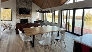 Un vaste espace de vie agrémenté d’une cheminée en pierre, d’une grande table à manger en bois et offrant une vue sur un lac grâce à de larges fenêtres.