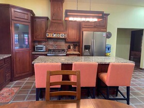 A kitchen with wooden cabinets, a stainless steel refrigerator, a stove, and a bar area with chairs. - Casa Serenidad | Pete's Camp | Baja 250 Rental | Sleeps 5 (San Felipe)