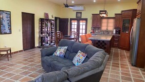 A living room with a sofa, a bookshelf, a dining area with chairs, and a kitchen with cabinets and a refrigerator. - Casa Serenidad | Pete's Camp | Baja 250 Rental | Sleeps 5 (San Felipe)