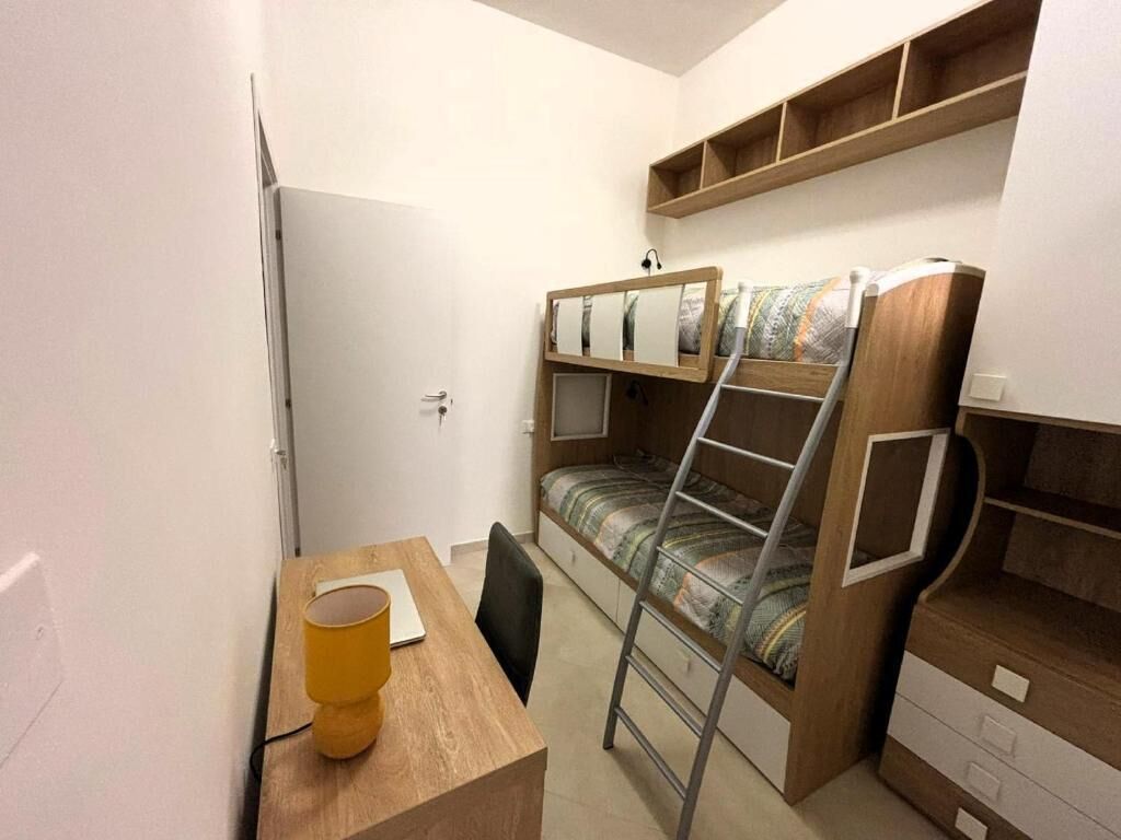 A compact room with a bunk bed, a desk with a chair, and a small shelf above the bed.