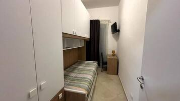 Una habitación pequeña y estrecha con una cama, un televisor en la pared y una puerta.