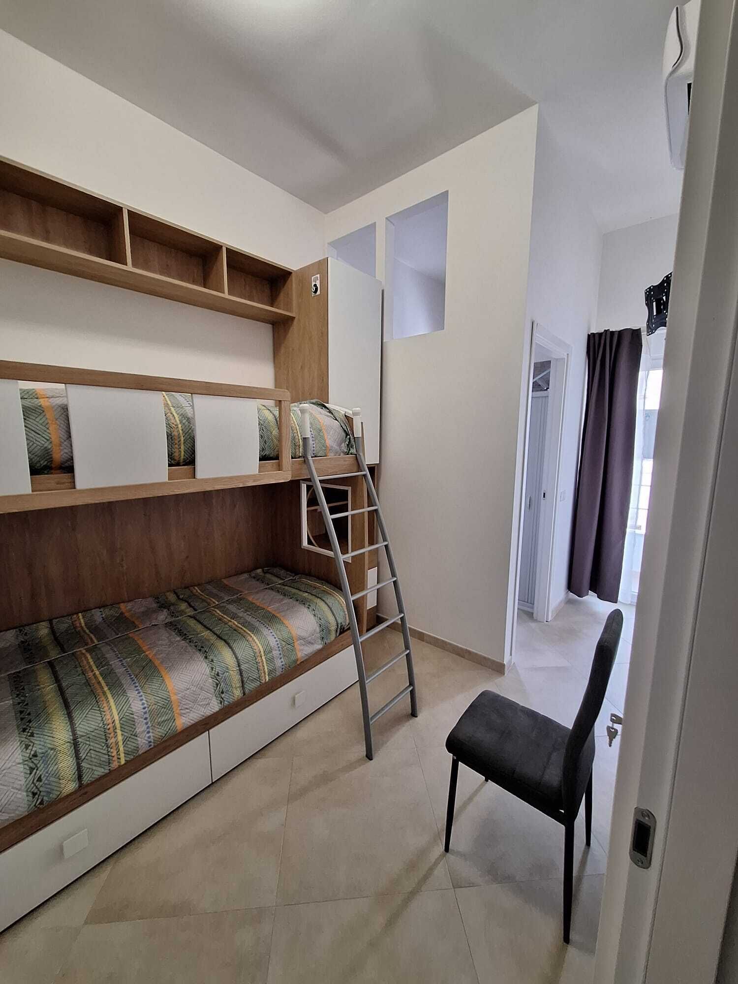 Habitación tipo dormitorio con literas, una escalera, una silla y una puerta que da a otra habitación.