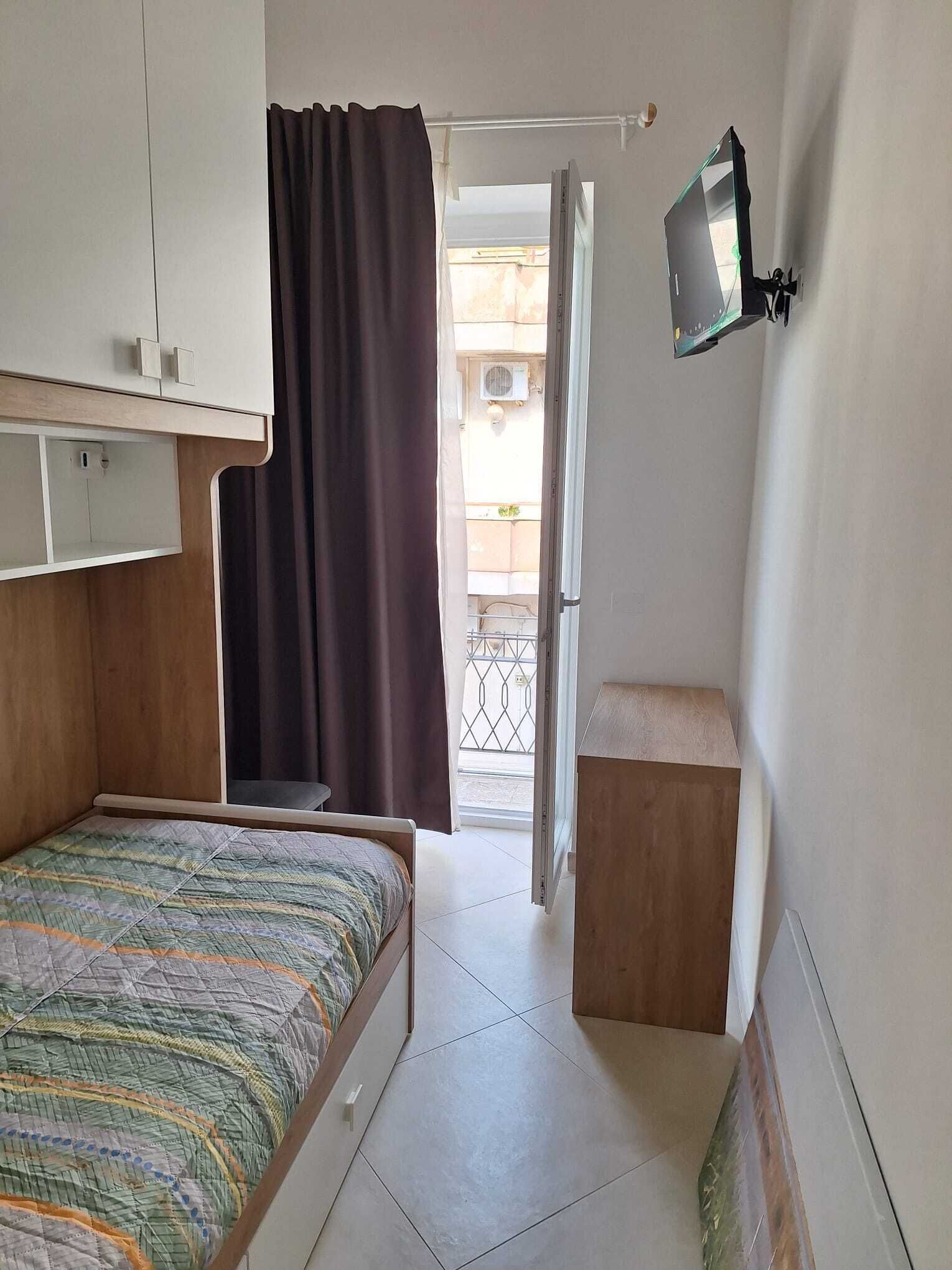 A compact room with a bed, a small table, a TV mounted on the wall, and a window with a curtain.