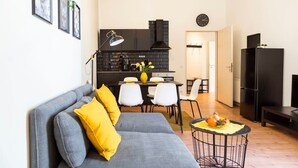Een moderne woonkamer met een grijze bank, gele kussens, een eethoek met witte stoelen, een zwarte keuken met een fornuis en een zwarte koelkast.