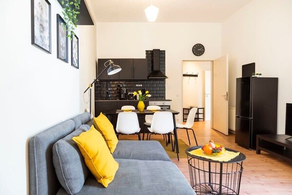 A modern living room with a grey sofa, yellow cushions, a dining area with white chairs, a black kitchen with a stove, and a black refrigerator.