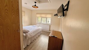 Een slaapkamer met een bed, een houten kledingkast, een aan de muur bevestigde televisie, een raam met jaloezieën en een plafondlamp.