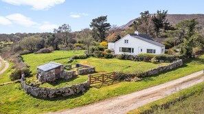 A white house with a stone wall and a wooden gate, surrounded by a green field and trees.