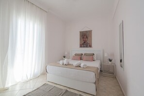 A bedroom with a bed, a bedside table, a nightstand, a painting, and a window with curtains. - MelMare Azuro Apartment by RentalsPro (Nea Moudania)