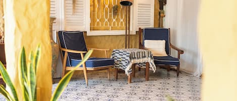 A room with two chairs, a small table, patterned floor tiles, and a decorative window with shutters.