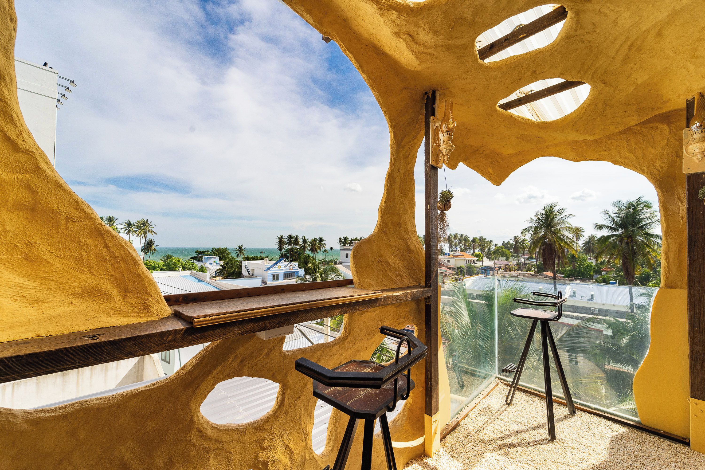 Ein Zimmer mit außergewöhnlichem architektonischen Design, das große, runde Ausschnitte und einen Balkon mit Blick auf eine Küstenlandschaft mit Palmen und Meerblick aufweist.