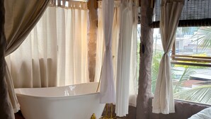 Une salle de bain avec une baignoire sur pieds, des rideaux blancs et une vue sur les palmiers à l’extérieur.