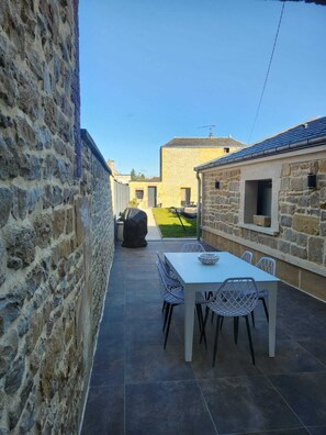 Un edificio de piedra con un patio que incluye una mesa, sillas, una parrilla y vistas a un césped verde y otro edificio en la lejanía.