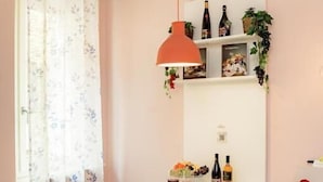 A dining area with a table, chairs, a hanging pendant light, a window with floral curtains, and a shelf with bottles and plants. - LA CASA DEL VINO-THE WINE HOUSE (Parma)