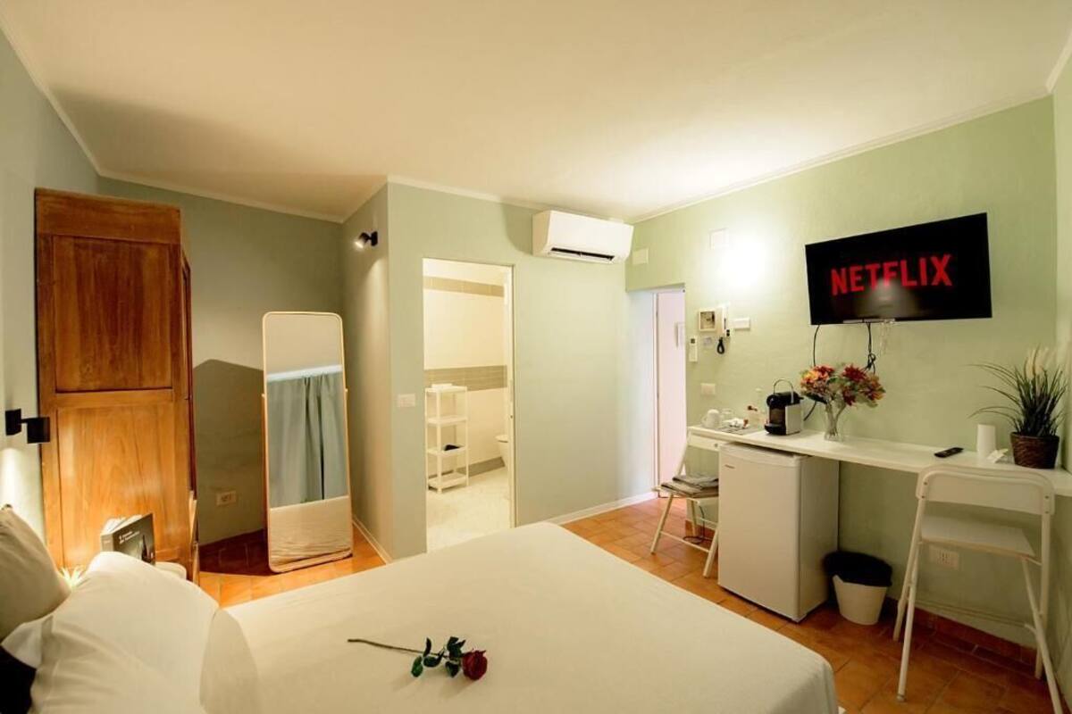 Una habitación de hotel con una cama, un escritorio con una computadora, un televisor mostrando 'NETFLIX' y una kitchenette pequeña.