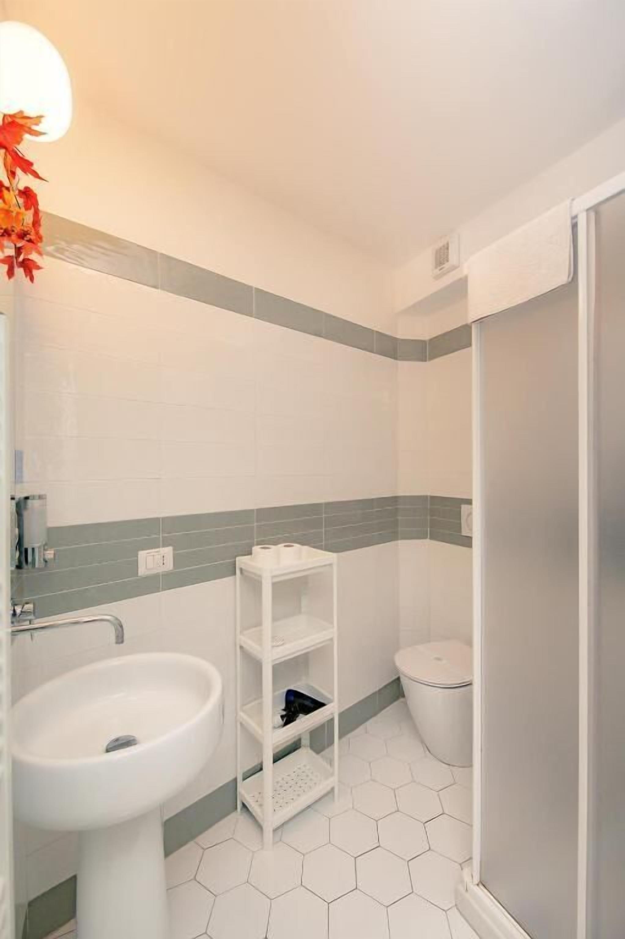 Un baño con lavamanos blanco, inodoro, ducha y repisa con artículos de aseo.