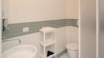 Un baño con lavamanos blanco, inodoro, ducha y repisa con artículos de aseo.