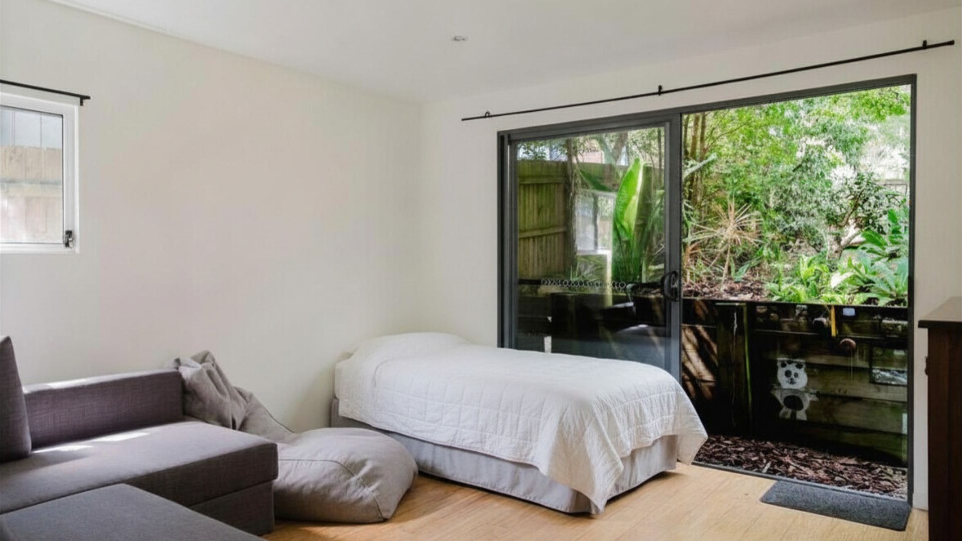 A bedroom with a bed, a sofa, and a sliding glass door leading to a balcony with plants.