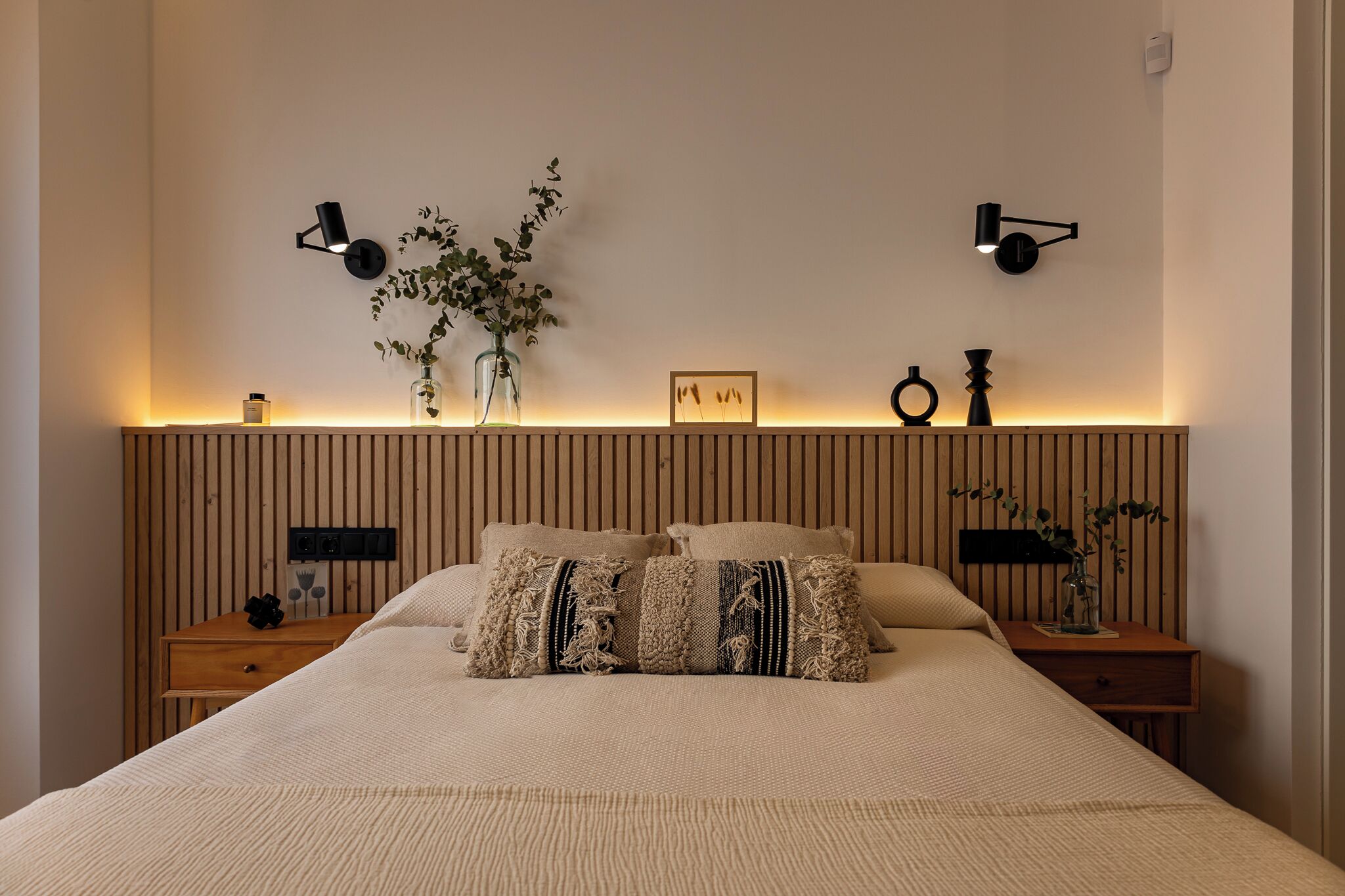 Un dormitorio con cama, mesitas de noche, lámparas de pared y objetos decorativos.