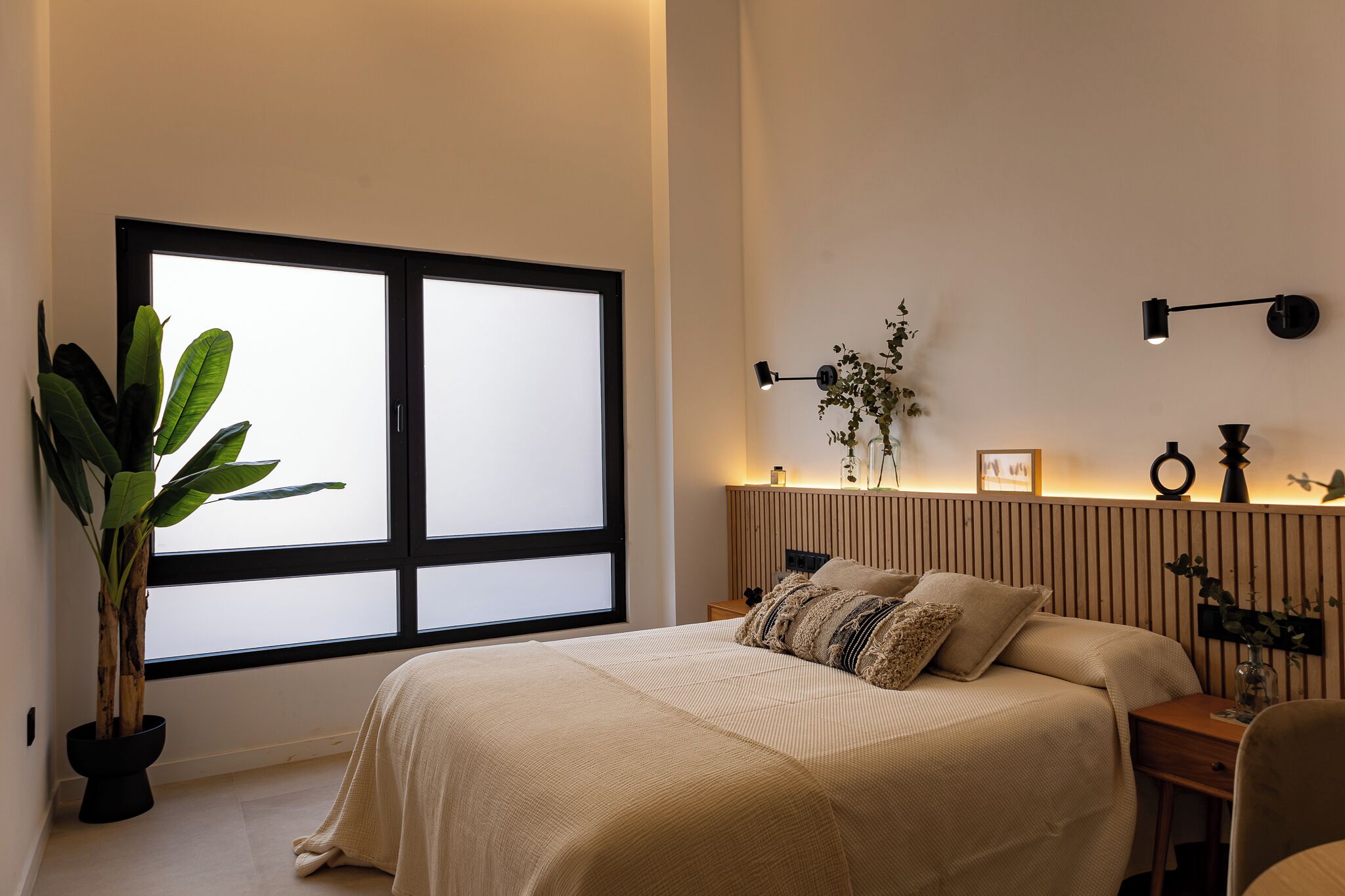 A bedroom with a large window, a bedside table with a lamp, a potted plant, and a bed with a beige blanket.