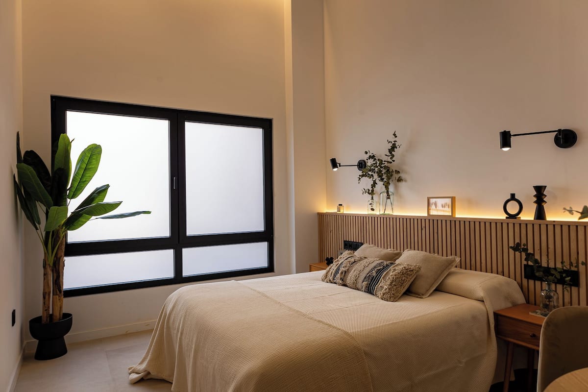 A bedroom with a large window, a bedside table with a lamp, a potted plant, and a bed with a beige blanket.