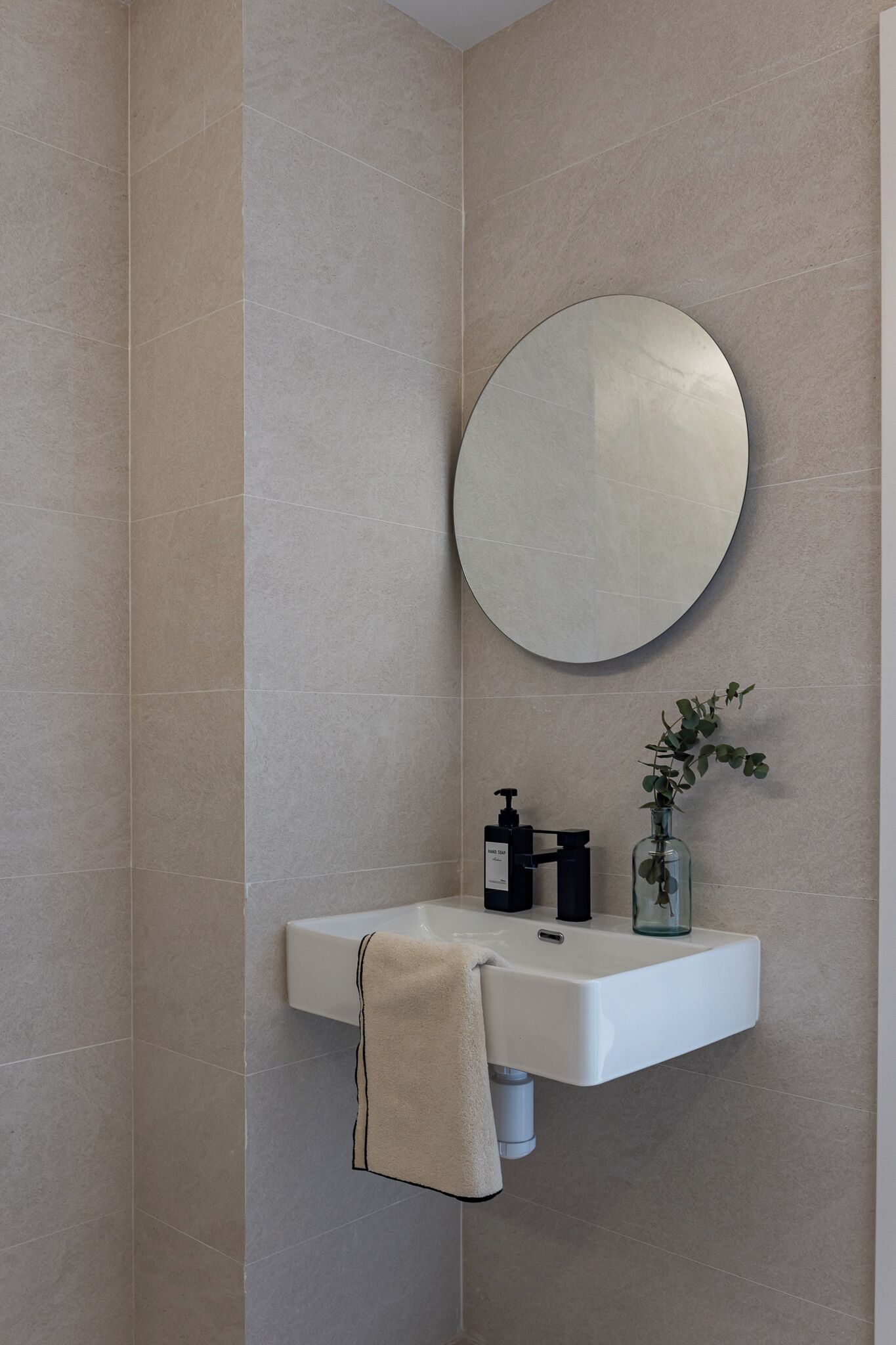 A bathroom with a white sink, a round mirror, a towel hanging on the wall, and a glass bottle with a plant.