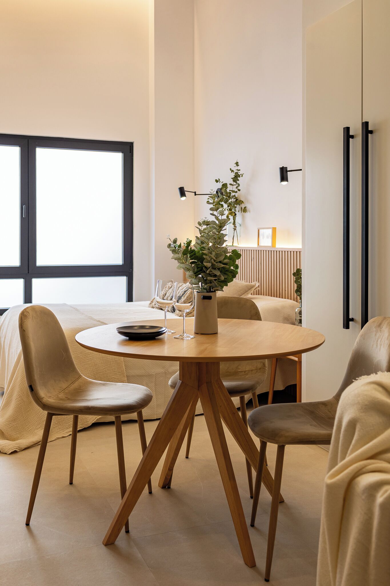 A modern hotel room with a round dining table, four chairs, a potted plant, and a bed with a large window.