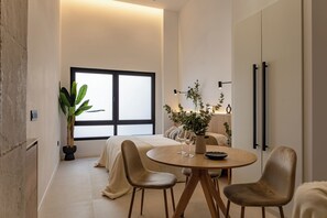 A modern bedroom with a large window, a bedside table with a plant, and a round dining table with chairs. - Almirante Suites  (València)