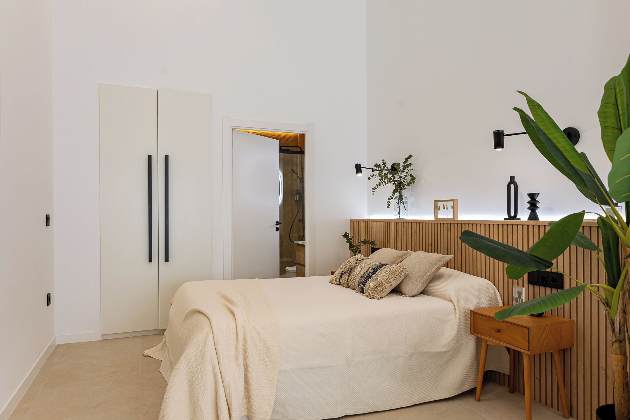 A bedroom with a bed, bedside table, lamp, and a plant.