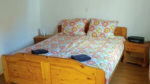 A wooden bed with a colorful floral bedspread, a bedside table with a lamp, and a small wooden nightstand with a radio.