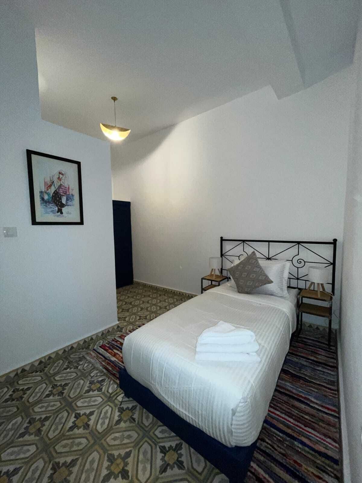 Un dormitorio con cama, cabecero, mesita de noche, lámpara, un cuadro enmarcado en la pared y alfombra estampada.