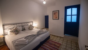 Un dormitorio con una cama, mesitas de noche, una alfombra estampada, una puerta azul y un cuadro en la pared.