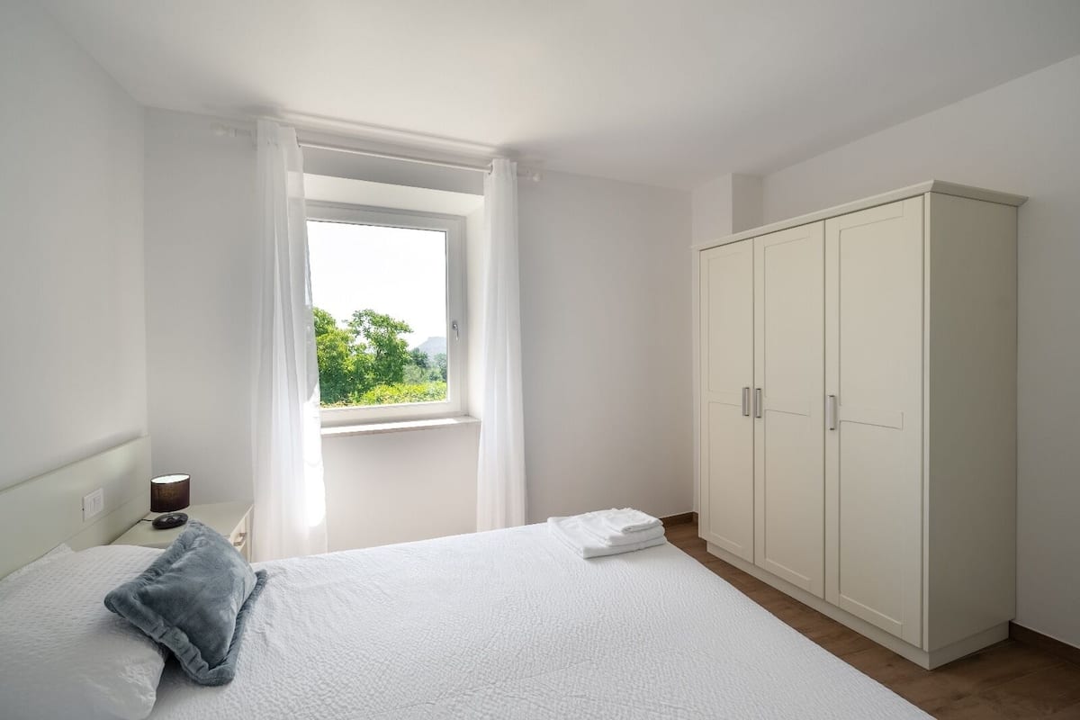 A bedroom with a bed, a window with curtains, a wardrobe, and a lamp on a bedside table.